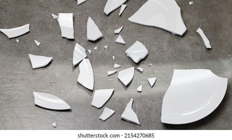 Closeup of broken dishes and white shards on the gray floor tiles background, splinters of break plate. Fragments tableware in kitchen. Shard, pieces of shattered dish ceramic. top view