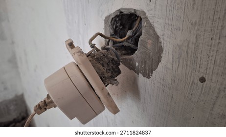 Close-up of a broken electrical socket pulled out from a rough wall, exposing dangerous tangled wiring and a deep hole, symbolizing danger, repair, construction, or electrical failure.