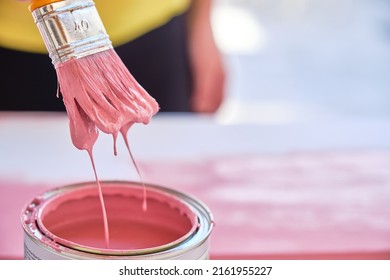 Close-up Of A Brush Dipping In A Pot Of Pink Paint Dripping With Boards And A Woman Out Of Focus In The Background