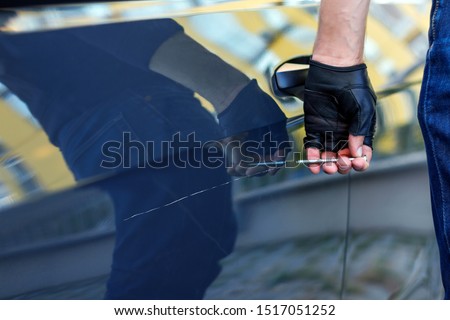 Closeup bully hands in black gloves are scratching automobile with nail, pin. Bandit is spoiling appearance of private car. Man is making scratch on auto without signaling. Vandalism act concept.