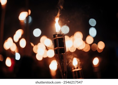 Close-up of a burning torch against a background of blurred lights, creating a bokeh effect. The flame dances gracefully, illuminating the night with a soft, warm glow - Powered by Shutterstock