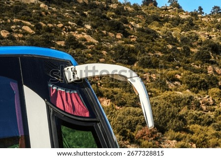 Close-up of bus side mirror with reflection in glass, red curtain and blue roof against rocky mountainside background. Concept of transport, travel, tourism, road trip and scenic journey in nature.