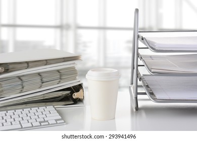 Closeup of a business desk with in-box-, binders, keyboard and disposable coffee cup in front of a large modern office window. The window is out of focus and high key. All items are white or silver. - Powered by Shutterstock
