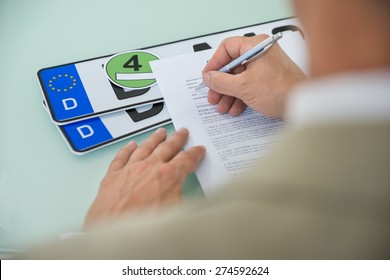 Close-up Of A Businessman Filling Car Sale Contract Form With Vehicle Registration Plate On Desk. Contract Paper Contains Placeholder Text