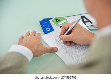 Close-up Of A Businessman Filling Car Sale Contract Form With Vehicle Registration Plate On Desk. Contract Paper Contains Placeholder Text