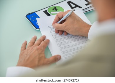 Close-up Of A Businessman Filling Car Sale Contract Form With Vehicle Registration Plate On Desk. Contract Paper Contains Placeholder Text