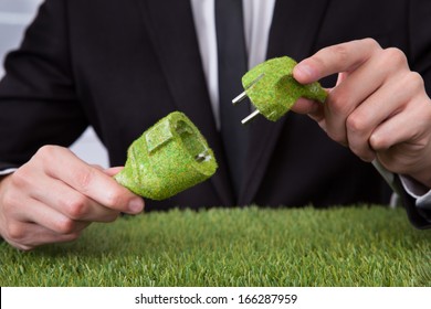 Close-up Of Businessman Hand Holding Grass Covered Electric Plug And Socket