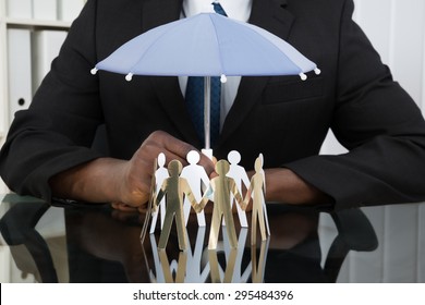 Close-up Of Businessman Hands Holding Small Umbrella Over Paper Cutout People At Desk - Powered by Shutterstock