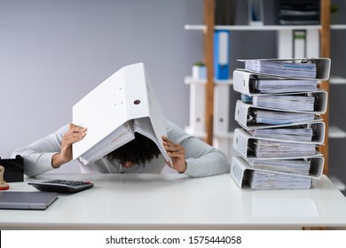 Close-up Of Businessman Hiding Under Folder At Desk With Stack Of Folders And Laptop