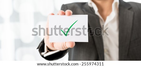 Closeup of businessman or teacher holding up a white card with green check mark. Conceptual of successfully completed task or homework.