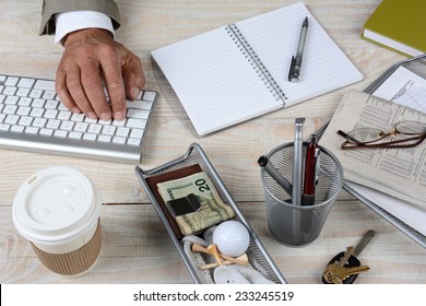 Closeup Of A Businessman's Desk. The White Rustic Wood Desk Has A Keyboard, Coffee Cup, Keys, Notebook, Glasses, Pencil Cup, Newspaper Money And Golf Ball And Tee, High Angle Shot With The Man Typing.