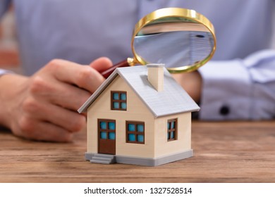 Close-up Of A Businessman's Hand Holding Magnifying Glass Over House Model Over Desk