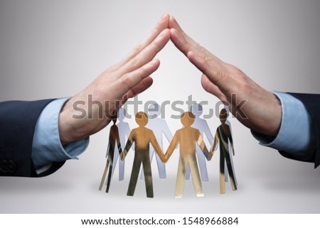 Close-up Of Businessman's Hand Protecting The Circle Of White Paper Cut-out Figures On Gray Background