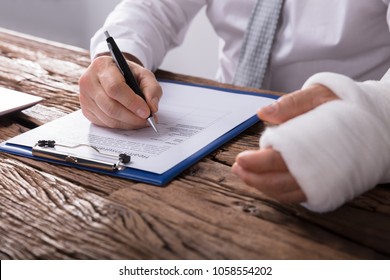 Close-up Of A Businessperson With Broken Arm Filling Health Insurance Claim Form On Wooden Desk