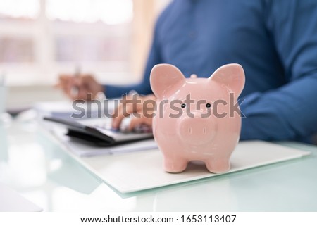 Close-up Of Businessperson With Piggybank Calculating Tax At Desk