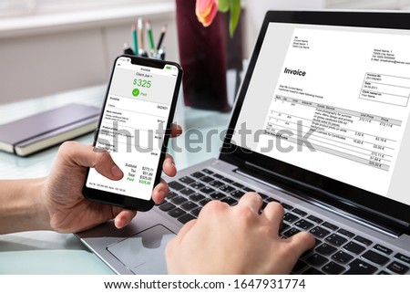 Close-up Of Businessperson Using Laptop While Paying Invoice On Mobilephone In Office
