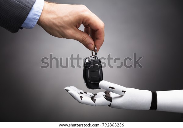Close-up Of A Businessperson\'s Hand Giving Car\
Key To Robot On Grey\
Background