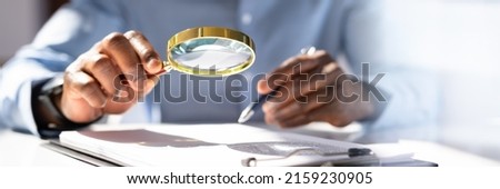 Close-up Of A Businessperson's Hand Holding Magnifying Glass Over Invoice At Workplace