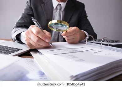 Close-up Of A Businessperson's Hand Looking At Receipts Through Magnifying Glass At Workplace