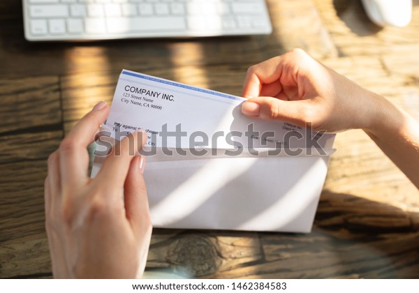 Close-up Of A Businessperson\'s Hand Opening\
Envelope With Paycheck Over Wooden\
Desk