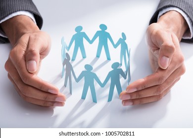 Close-up Of A Businessperson's Hand Protecting Cut-out Figures On Desk - Powered by Shutterstock