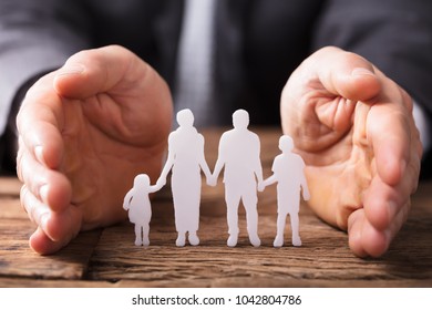 Close-up Of A Businessperson's Hand Protecting Family Figures On Wooden Desk - Powered by Shutterstock