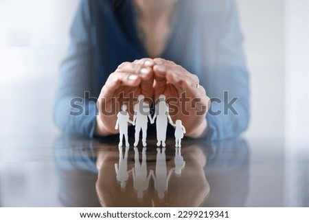 Close-up Of A Businessperson's Hand Protecting Family Figures On Wooden Desk