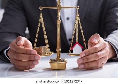 Close-up Of A Businessperson's Hand Protecting Stacked Coins And House Model On Weighing Scale