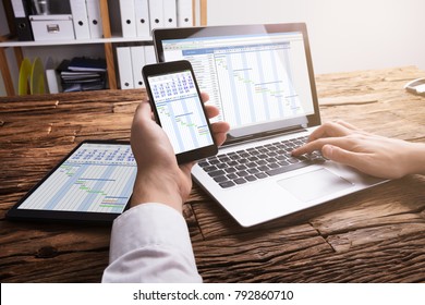 Close-up Of A Businessperson's Hand Using Smartphone While Analyzing Gantt Chart On Laptop