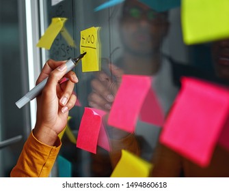 Close-up of businesswoman hand writing on yellow sticky notes stuck over glass board in creative office