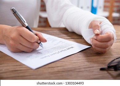 Close-up Of Businesswoman With White Bandage Hand Filling Work Injury Claim Form On Wooden Desk