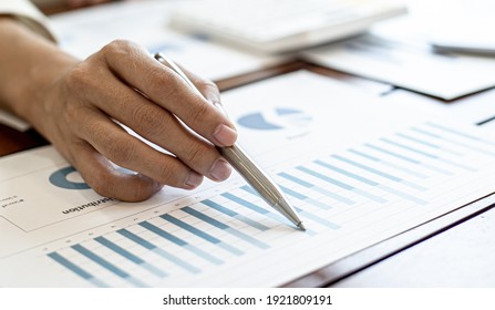 Close-up Businesswoman's Hand Holding A Pen Pointing At A Bar Chart On A Corporate Financial Information Sheet, The Businesswoman Examines The Financial Information Provided By The Finance Department.