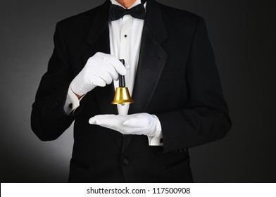 Closeup of a butler wearing a tuxedo and holding a service bell in front of his torso. Man is unrecognizable. Horizontal format on a light to dark gray background.