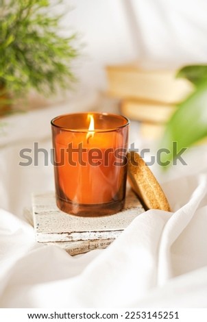 Closeup of candle in glass jar with blank label. Blurred green plant, white linen tablecloth backgound. Front view. Handmade ecological and vegan soy wax candles. Mock up, space for text