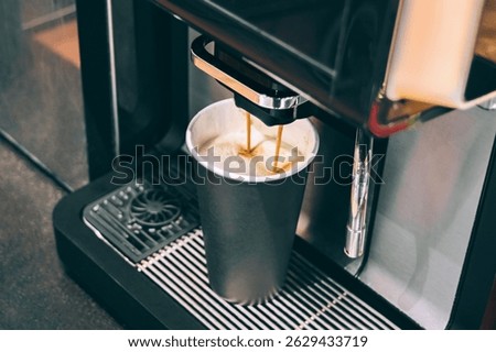 A close-up captures a coffee machine dispensing a stream of espresso into a tall black paper cup with a white interior, resting on the machine's metal drip grid. The focus is on the brewing process.