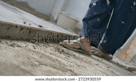 A close-up captures a construction worker's foot in a flip-flop resting on a dusty floor, highlighting work in progress. The image showcases the worker's leg and foot in casual attire amidst the construction site. Soft lighting illuminates the scene, offering space for editorial use.