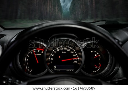 A close-up of a car panel with a speedometer indicating a huge speed of 200 km per hour, a tachometer at 6000 rpm, oil and gasoline levels. The car rides on the highway at high speed along the forest
