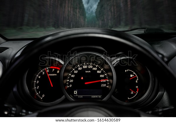A close-up of a car panel with a speedometer\
indicating a huge speed of 200 km per hour, a tachometer at 6000\
rpm, oil and gasoline levels. The car rides on the highway at high\
speed along the forest