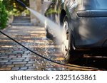 Close-up of car washing process. Unknown man directs a hose with jet of water at the rear wheel of his car, washes the vehicle in the backyard of the house. Car cleaning concept. People and transport