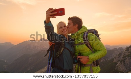 CLOSEUP: Carefree man kisses his smiling girlfriend taking a photo of them in the picturesque Alps. Adorable active couple enjoying a peaceful summer evening in the scenic mountains and taking photos.