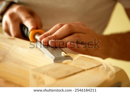 A close-up of a carpenter hands skillfully using a chisel on a wooden board. The focus is on the detailed work, with the wood grain visible and the chisel carving into the surface.
