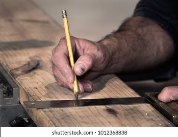 Closeup of carpenter's rough rugged hands and broken finger nails, using pencil and old wooden handle square to mark a line on a wood board to cut, with sawdust around