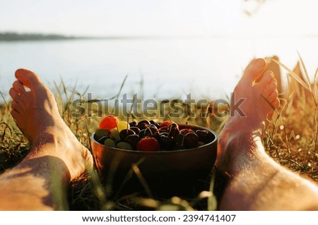 Close-up of caucasian bare male feet and plate of fruit on green grass on sunny summer day. Rest, relaxation in nature. Selective focus on cup with cherries, peach, strawberries, grapes.