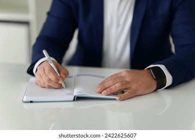 Close-up of caucasian businessman hands writing in a notebook, wearing a smartwatch and a blue suit, on a white table in a well-lit office setting, cropped. Work, business - Powered by Shutterstock