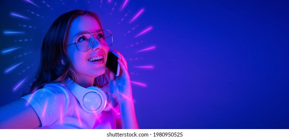 Close-up Caucasian girl's portrait isolated on blue background in multicolored neon light with luminescent rays. Beautiful model with phone. Concept of human emotions, facial expression, ad, fashion.