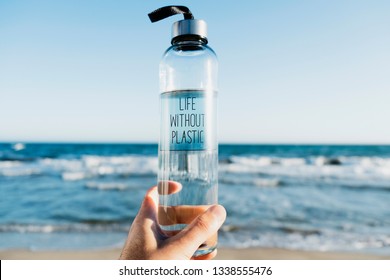closeup of a caucasian man holding a glass reusable water bottle with the text life without plastic written in it, on the beach, with the ocean in the background
