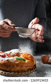 closeup of a caucasian man sprinkling some icing sugar on a roscon de reyes, a spanish three kings cake eaten on epiphany day, placed on a gray rustic table