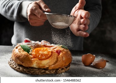 closeup of a caucasian man sprinkling some icing sugar on a roscon de reyes, a spanish three kings cake eaten on epiphany day, placed on a gray rustic table