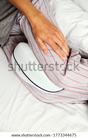 closeup of a caucasian man, wearing striped pajama pants, using an anatomical cushion between his legs while is lying on a bed set with white bedsheets
