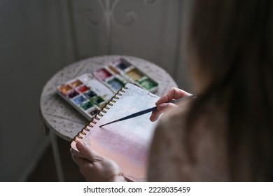 closeup of a caucasian mature woman's left hand holding a notebook while painting on it with a fine brush - Shutterstock ID 2228035459